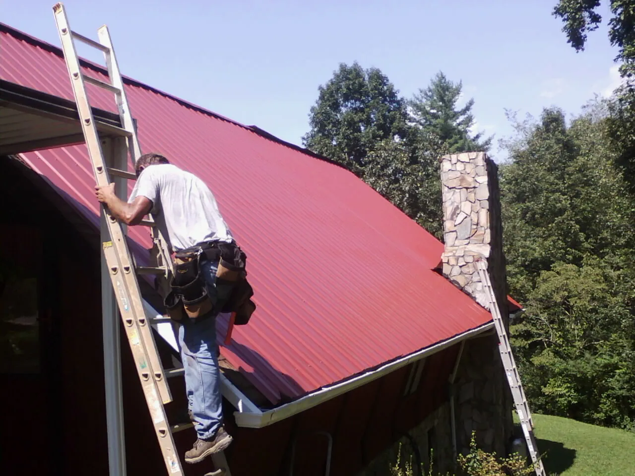 Roofer on a ladder beside a red metal roof and stone chimney — Soffit & Fascia Repair by Roofing Brothers in Roxbury